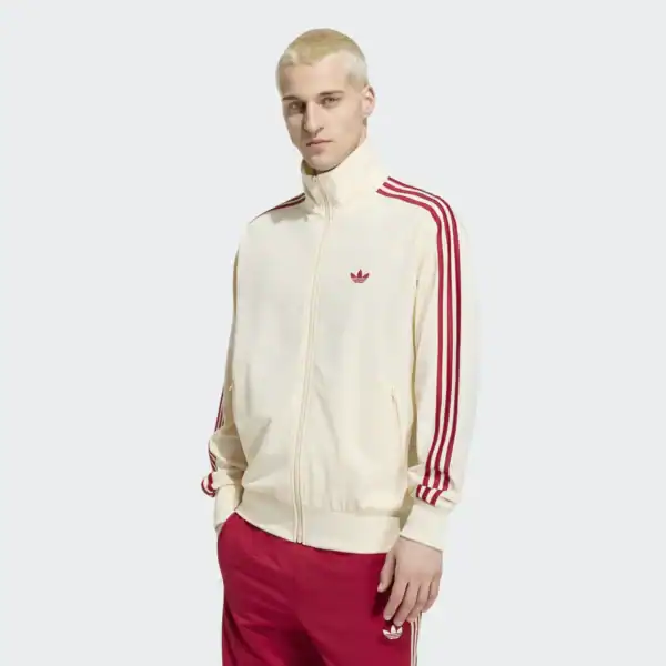 Mann trägt eine beige Adidas Firebird Originals Track Jacke mit roten Streifen und Logo, kombiniert mit roten Hosen, auf weißem Hintergrund.