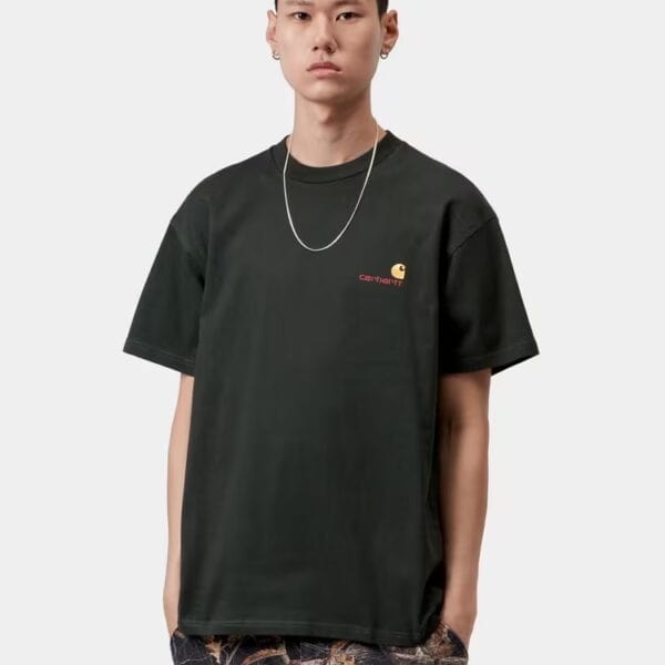 Junge Frau trägt ein dunkles Carhartt Wip T-Shirt in Raven grün mit Logo-Details, kombiniert mit Camouflage-Hose, auf einem neutralen Hintergrund.