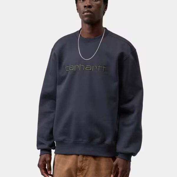Mann trägt einen dunklen Carhartt Wip Sweatshirt mit Logo-Print, kombiniert mit braunen Hosen, auf weißem Hintergrund.