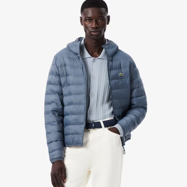 Mann trägt eine graue, wasserdichte Daunenjacke von Lacoste, perfekt für kalte und nasse Wetterbedingungen, kombiniert mit einem hellblauen Hemd und weißen Hosen.